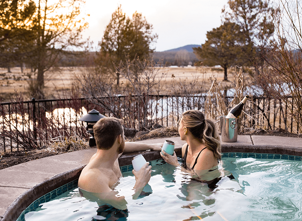 A man and woman relax in an outdoor hot tub, holding drinks and looking at the sunset over a scenic landscape with trees, grass, and a pond. A wine bucket sits on the tubs edge.