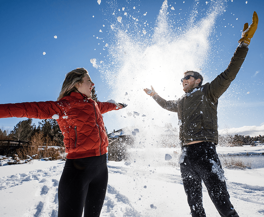 Two people in winter jackets laugh and throw snow up into the air on a sunny, snowy day. Snow is midair between them, and trees and blue sky are in the background.