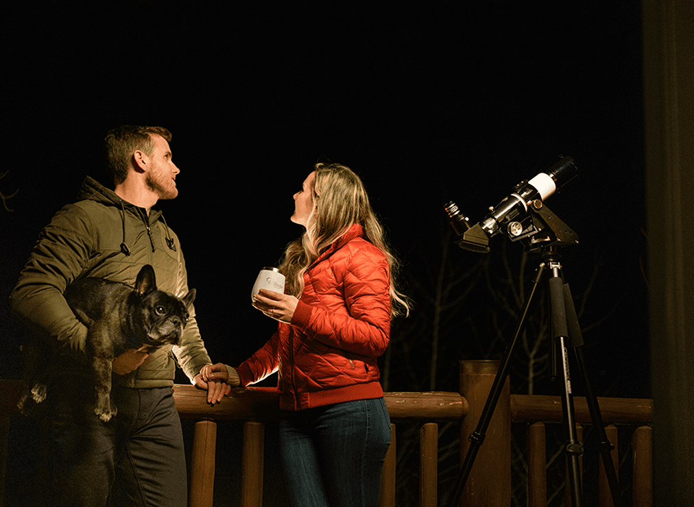 A man holding a dog and a woman with a mug stand on a wooden deck at night, looking up at the sky next to a telescope.