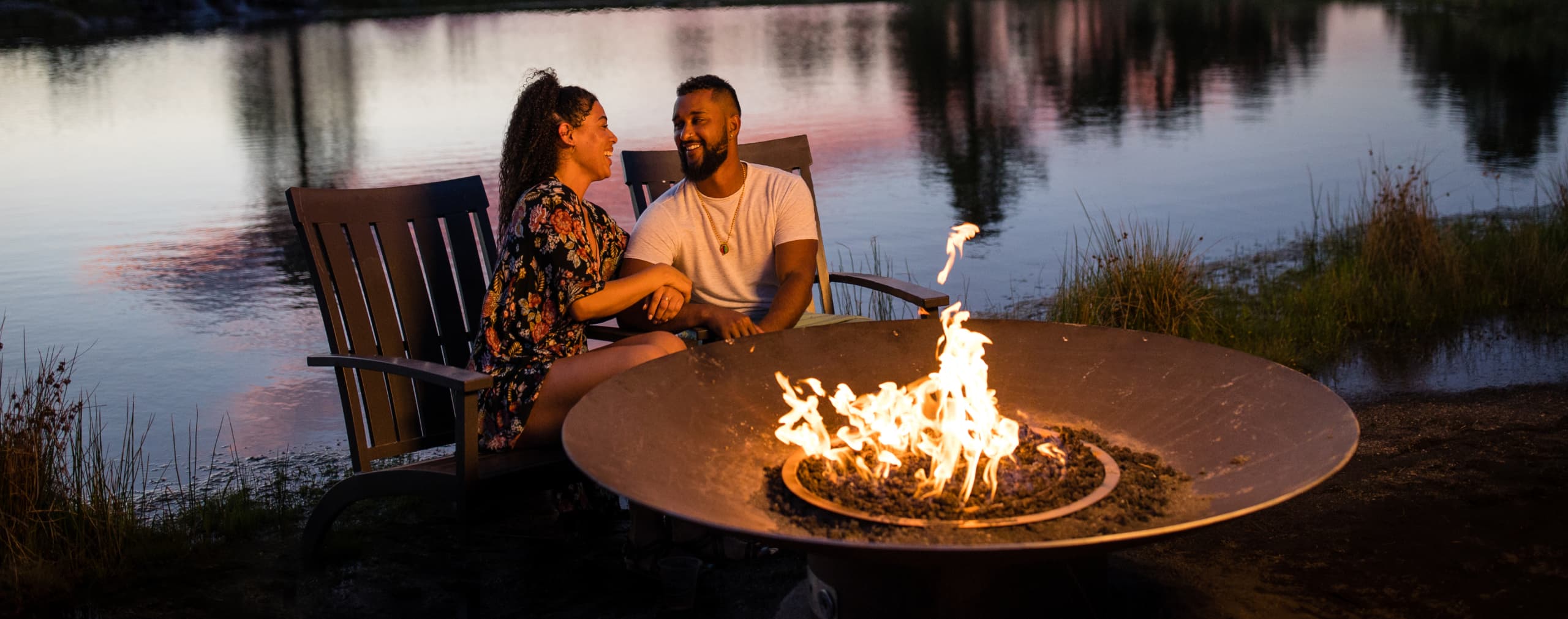 A couple sits together on wooden chairs by a lake at sunset, smiling at each other with a fire pit burning in front of them.