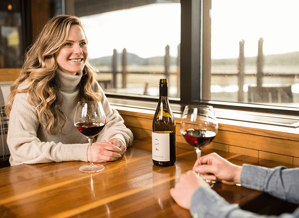 Two people sit at a wooden table by a window, enjoying glasses of red wine. One person smiles, while the other is partially visible, with a wine bottle in the center. Sunlight streams in through the window.