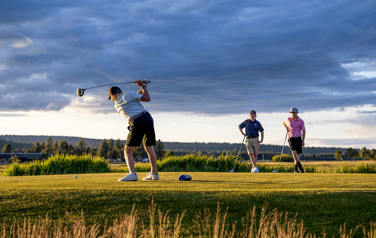 A golfer swings a club on a green course at sunset while two people watch in the background under a partly cloudy sky.