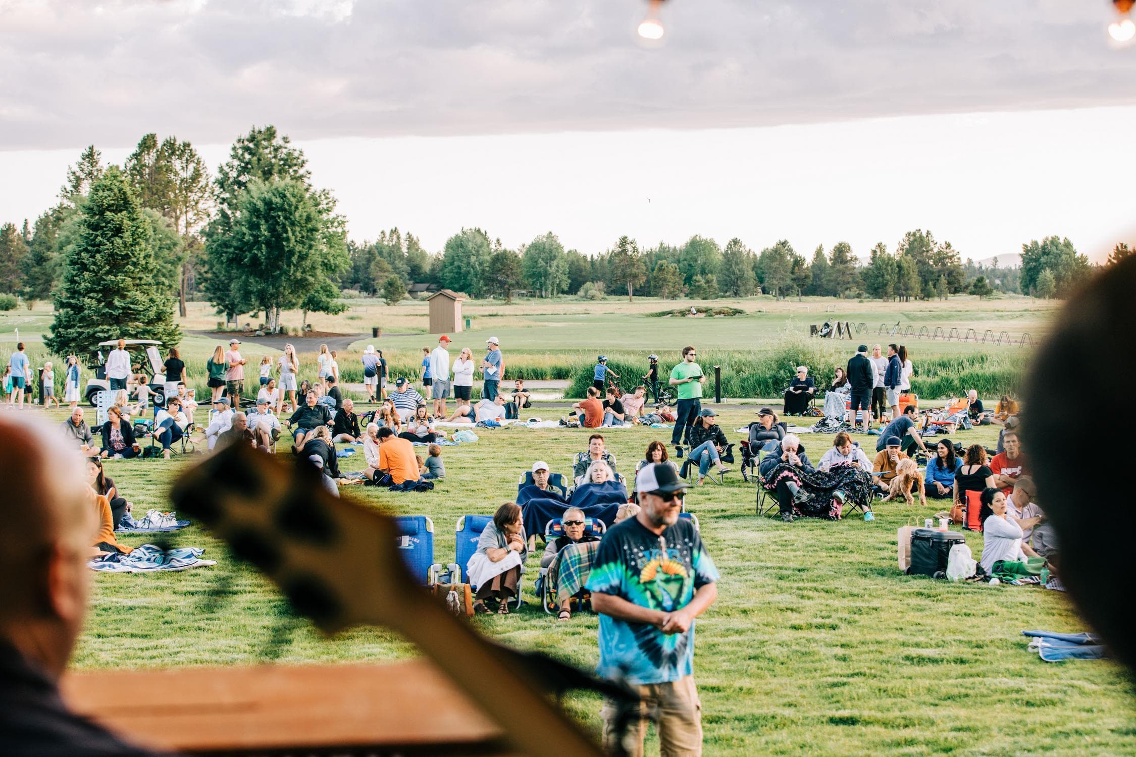 People gather outdoors on a grassy field, sitting on blankets and chairs, enjoying a community event under a cloudy sky. Some trees and a small building are visible in the background.