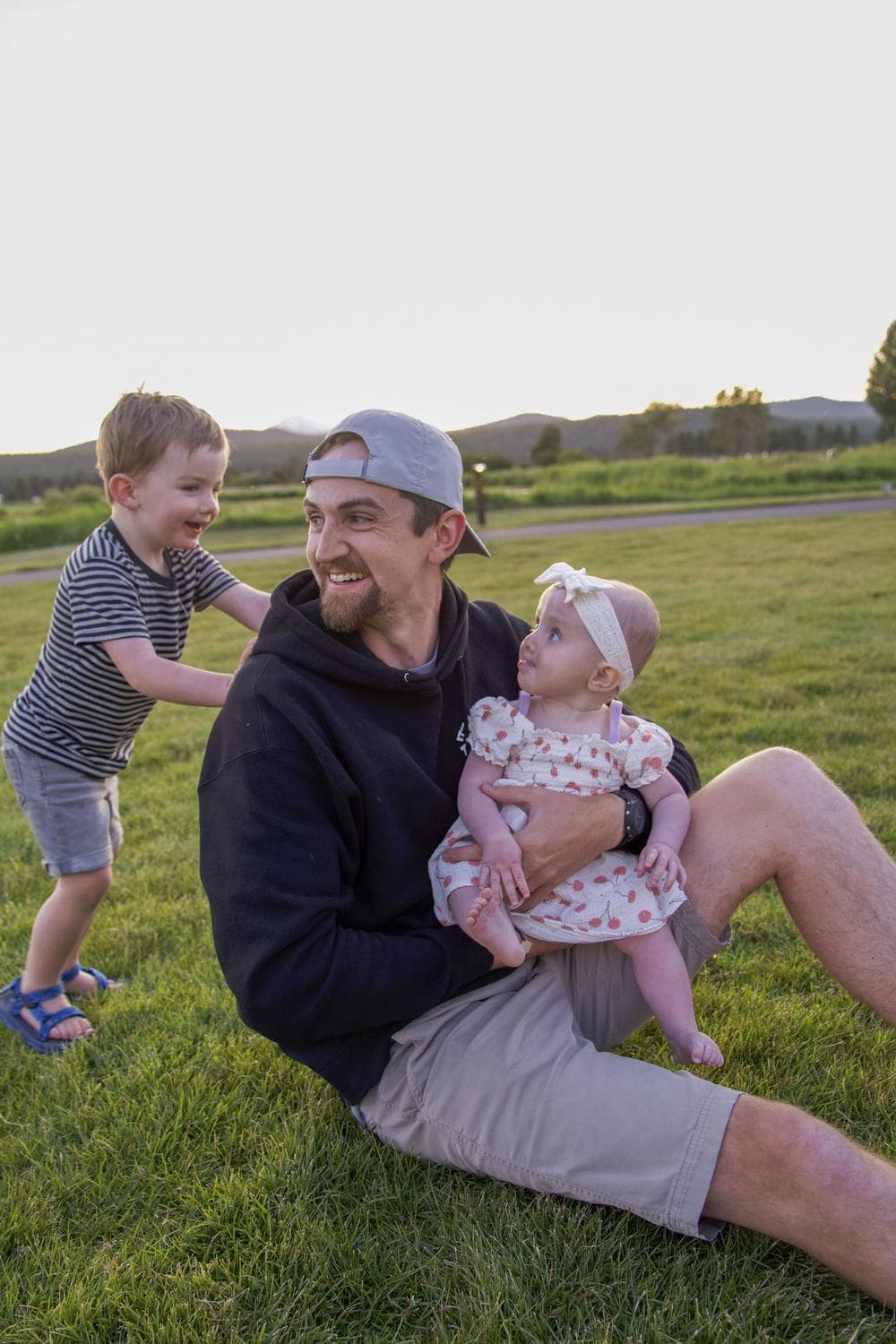 A man in a cap sits on grass holding a baby in a dress while a smiling boy in stripes playfully pushes him from behind.