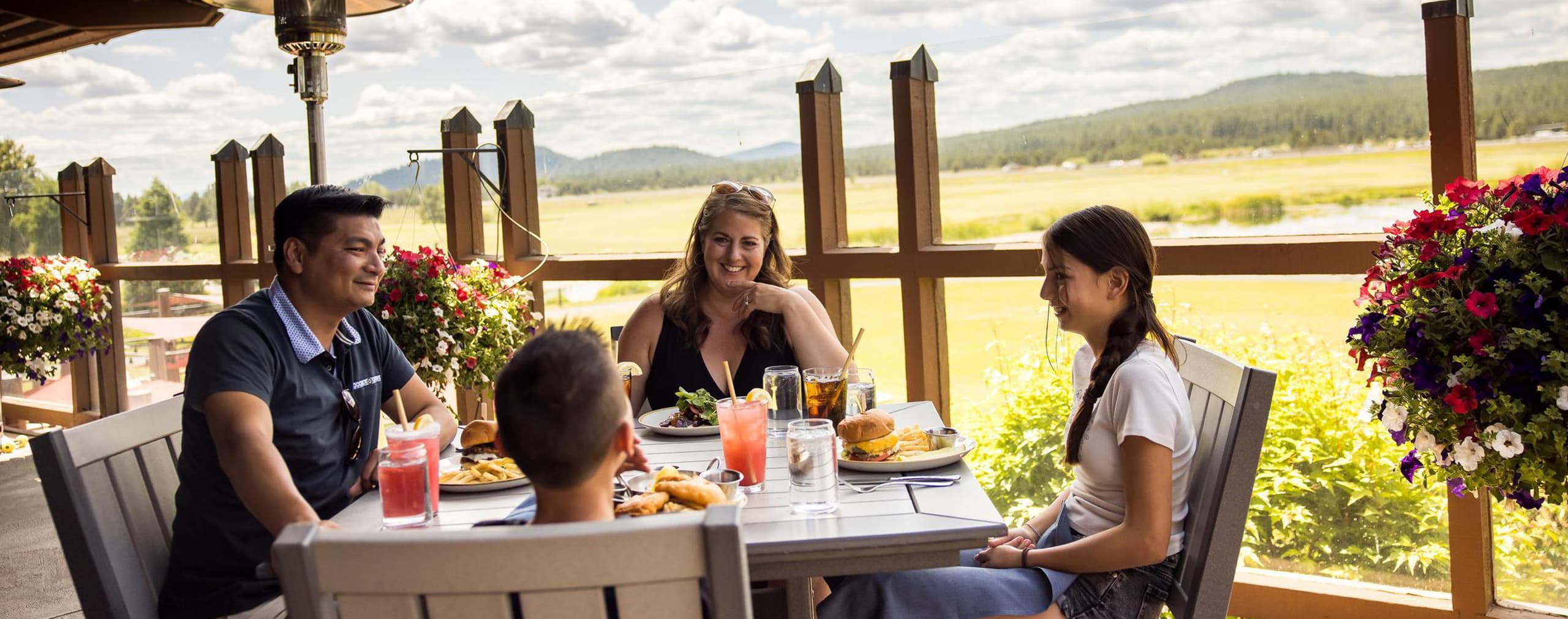A family of four dines outdoors on a sunny day, seated at a table with food and drinks. Colorful flowers and a scenic view of fields and trees are visible in the background.
