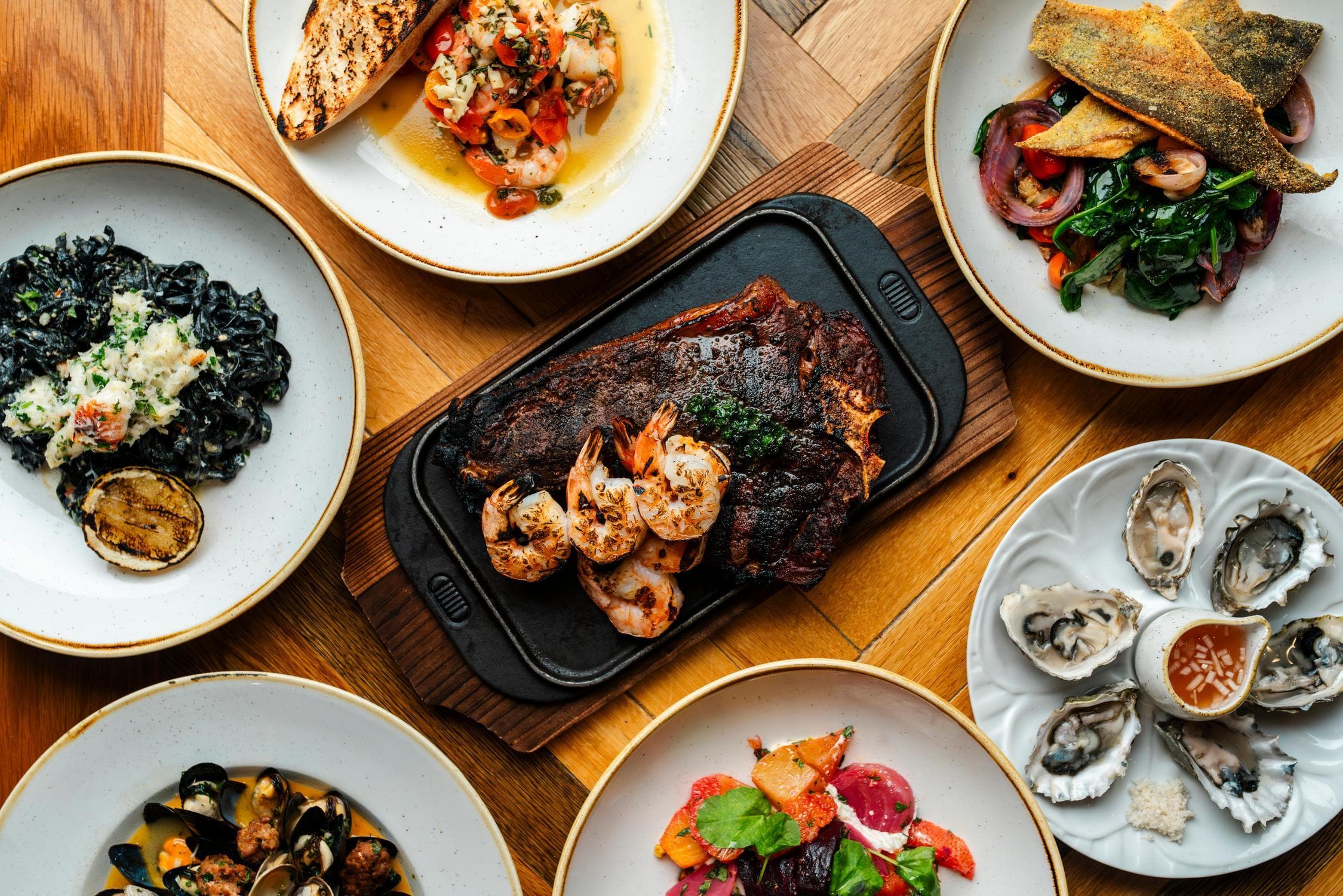 A table set with grilled steak and shrimp, oysters, black pasta, fish filet, seafood salad, and a beet salad, all served on white plates over a wooden surface.