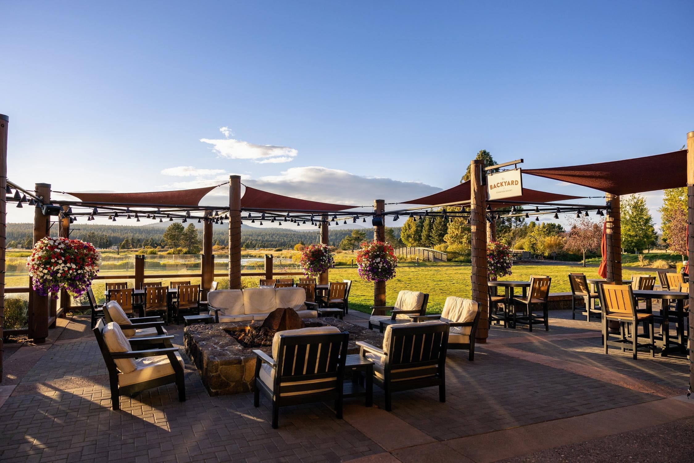 Outdoor patio with lounge chairs and tables around a central fire pit, overlooking a scenic grassy field and trees under a clear blue sky. Hanging flower baskets and sunshades add to the inviting atmosphere.