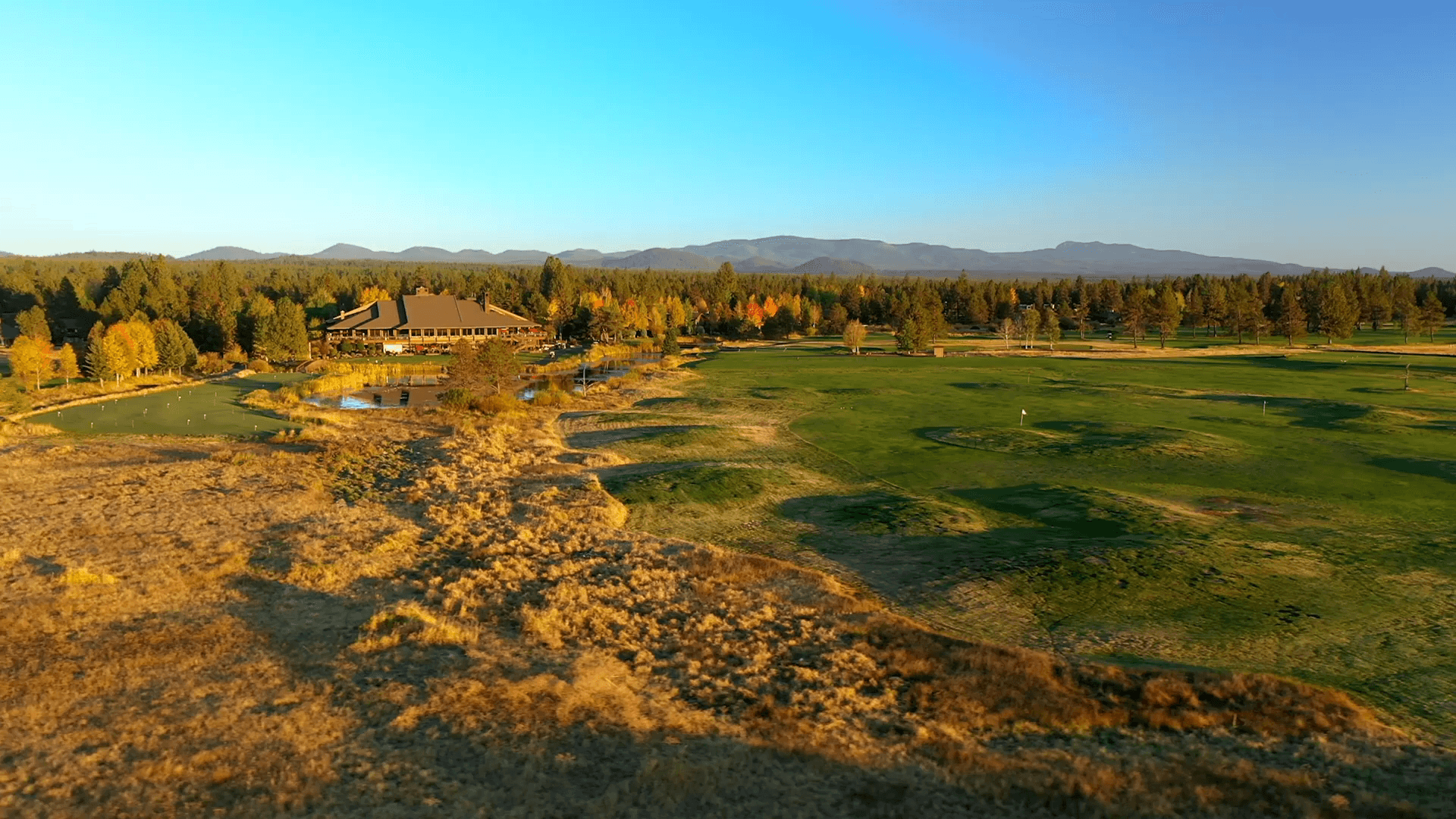 Aerial view of a golf course with a clubhouse surrounded by trees and mountains in the background under a clear blue sky.