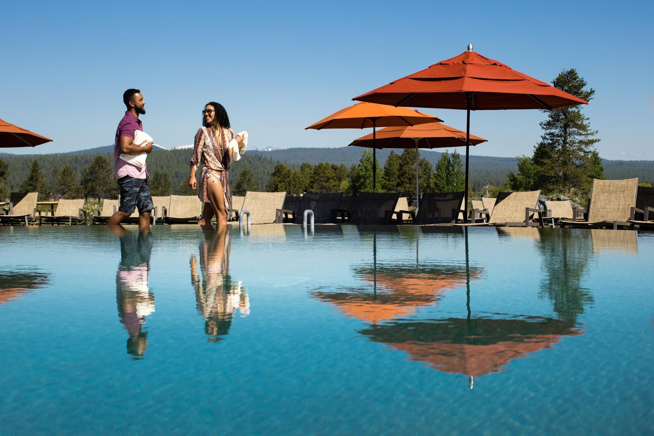 A man and a woman walk by a pool with orange umbrellas, lounge chairs, and trees in the background under a clear blue sky.