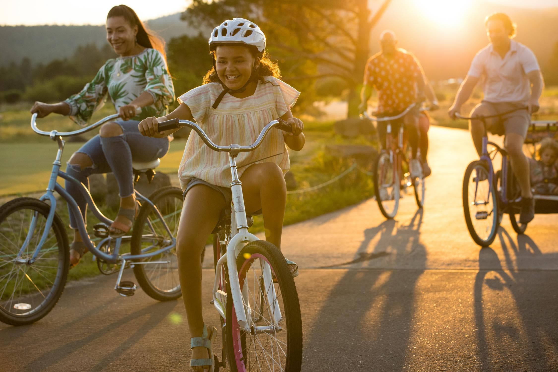 A group of people, including children and adults, ride bicycles together on a sunlit path in the late afternoon, smiling and enjoying the outdoors.
