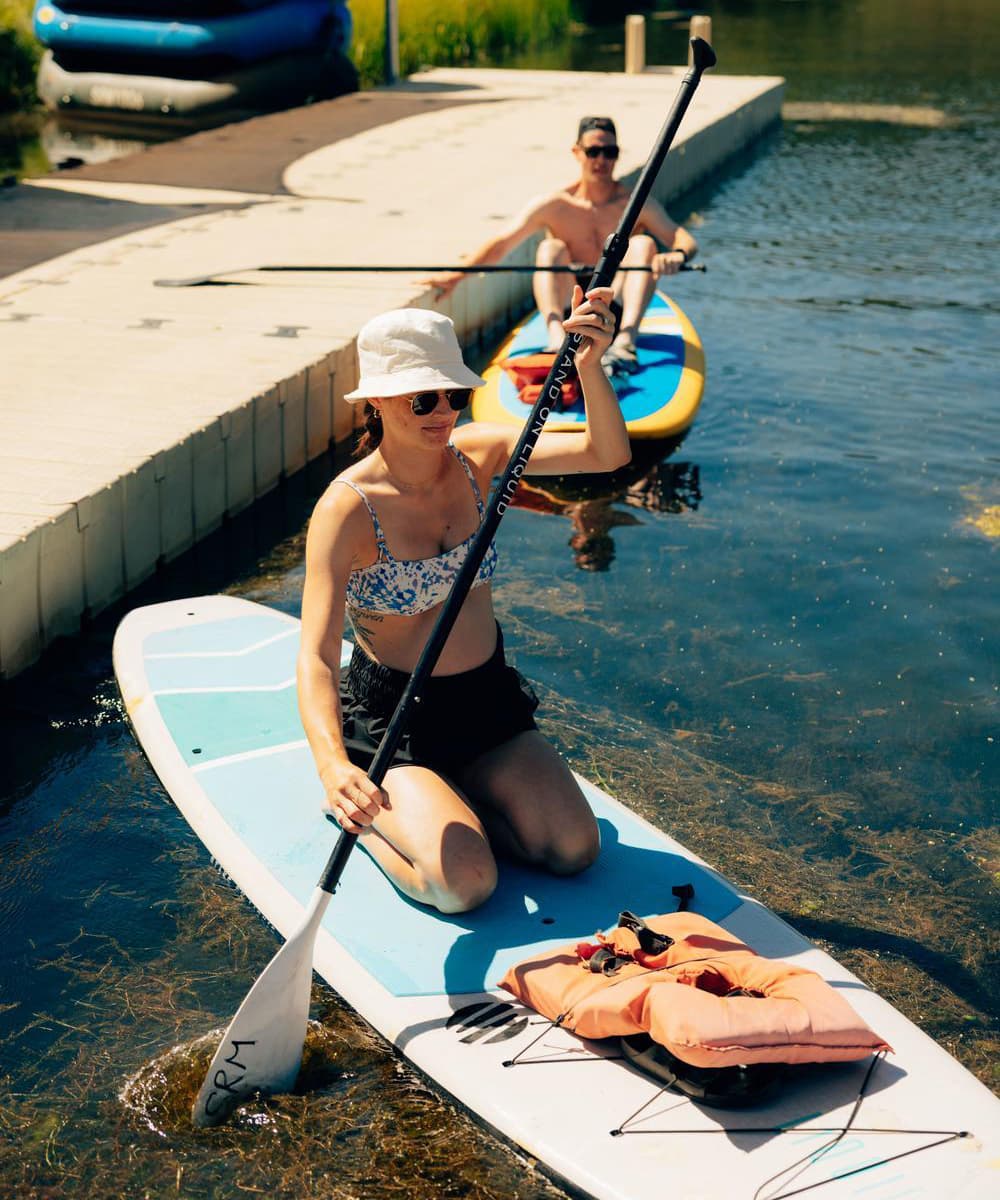 A person kneels on a paddleboard holding a paddle, wearing a sun hat and sunglasses. Another person sits in a kayak behind them, both on a sunny day by the dock.