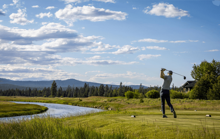 A golfer finishes a swing on a lush green course near a winding river, with trees and mountains under a partly cloudy sky.