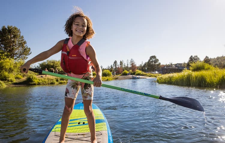 Smiling child wearing a red life jacket stands on a paddleboard, holding a paddle on a calm river with green grass and trees under a clear blue sky.