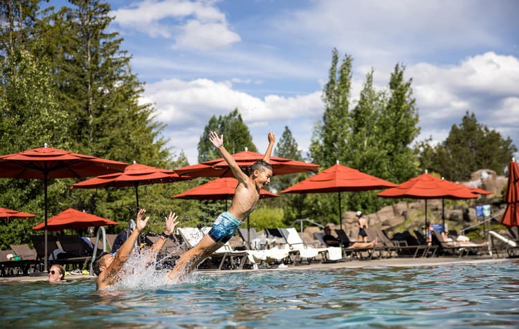 A child in swim trunks is playfully thrown into the air above a pool, arms outstretched, while adults watch nearby. Red umbrellas and lounge chairs are in the background.
