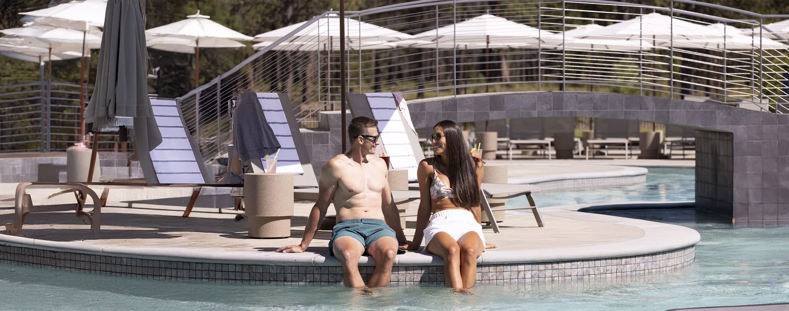 A man and woman sit at the edge of a pool with their feet in the water, talking and smiling. Lounge chairs, umbrellas, and a curved bridge are in the background.
