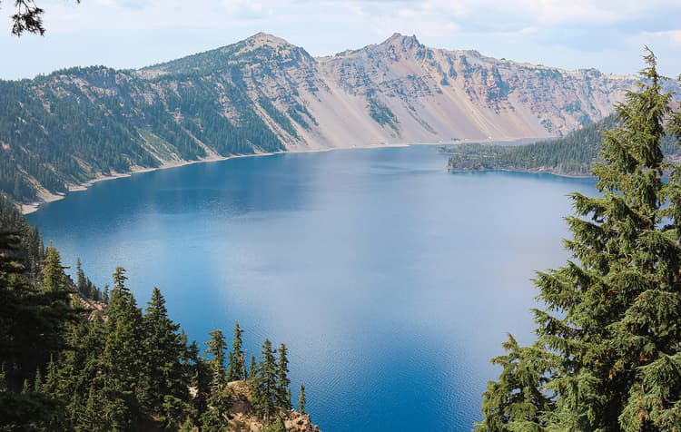 A deep blue lake surrounded by evergreen trees and steep, rocky mountain slopes under a clear sky.