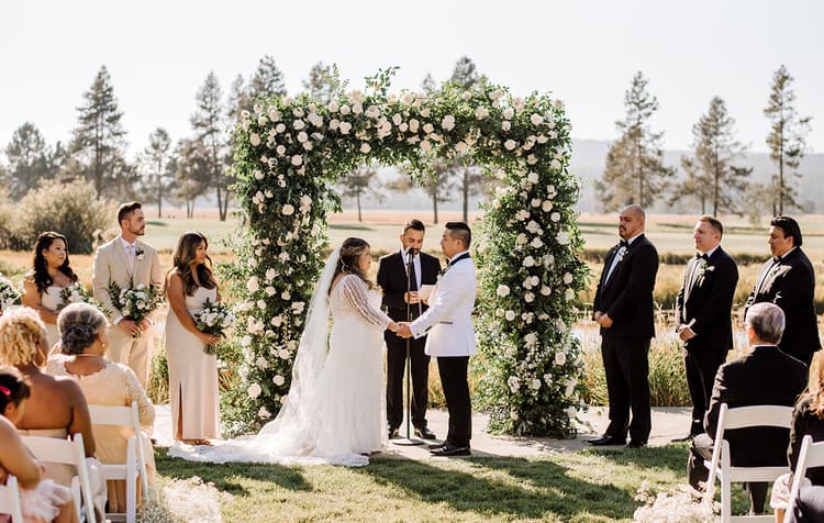 A couple stands under a floral arch, holding hands and facing each other during an outdoor wedding ceremony, surrounded by their wedding party and guests.