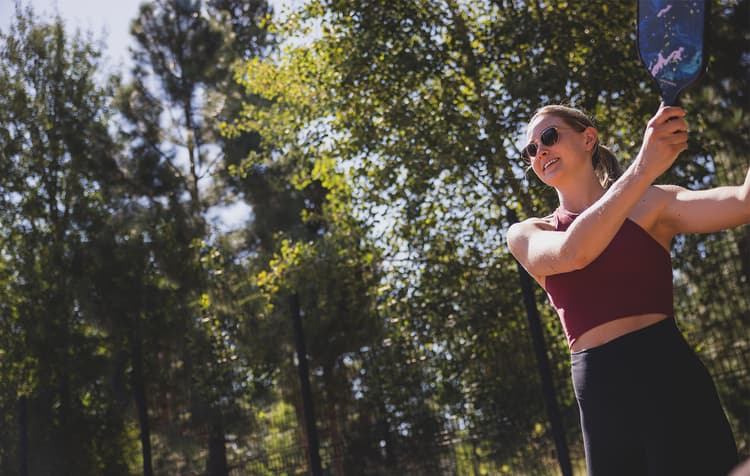 A woman in sunglasses and athletic clothes smiles while playing racket sports outdoors on a sunny day, surrounded by tall green trees.
