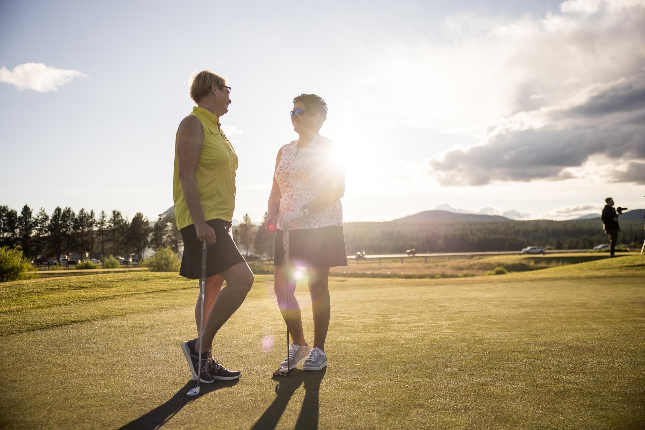Two women stand on a golf course at sunset, talking and smiling. Trees, mountains, and another golfer are visible in the background with sunlight shining behind them.
