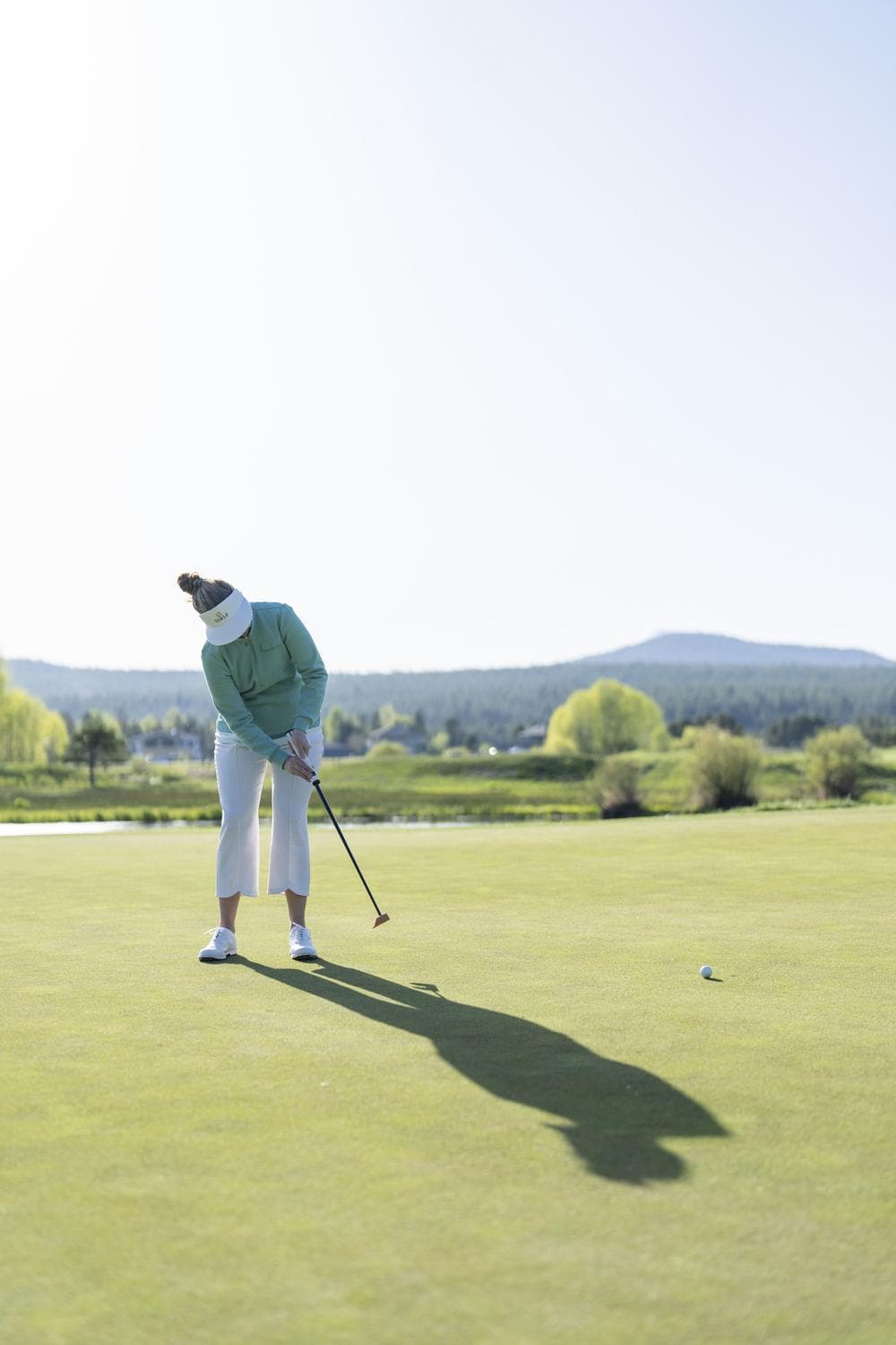 A person wearing a visor and light clothing is putting a golf ball on a sunny green, with mountains and trees in the background.