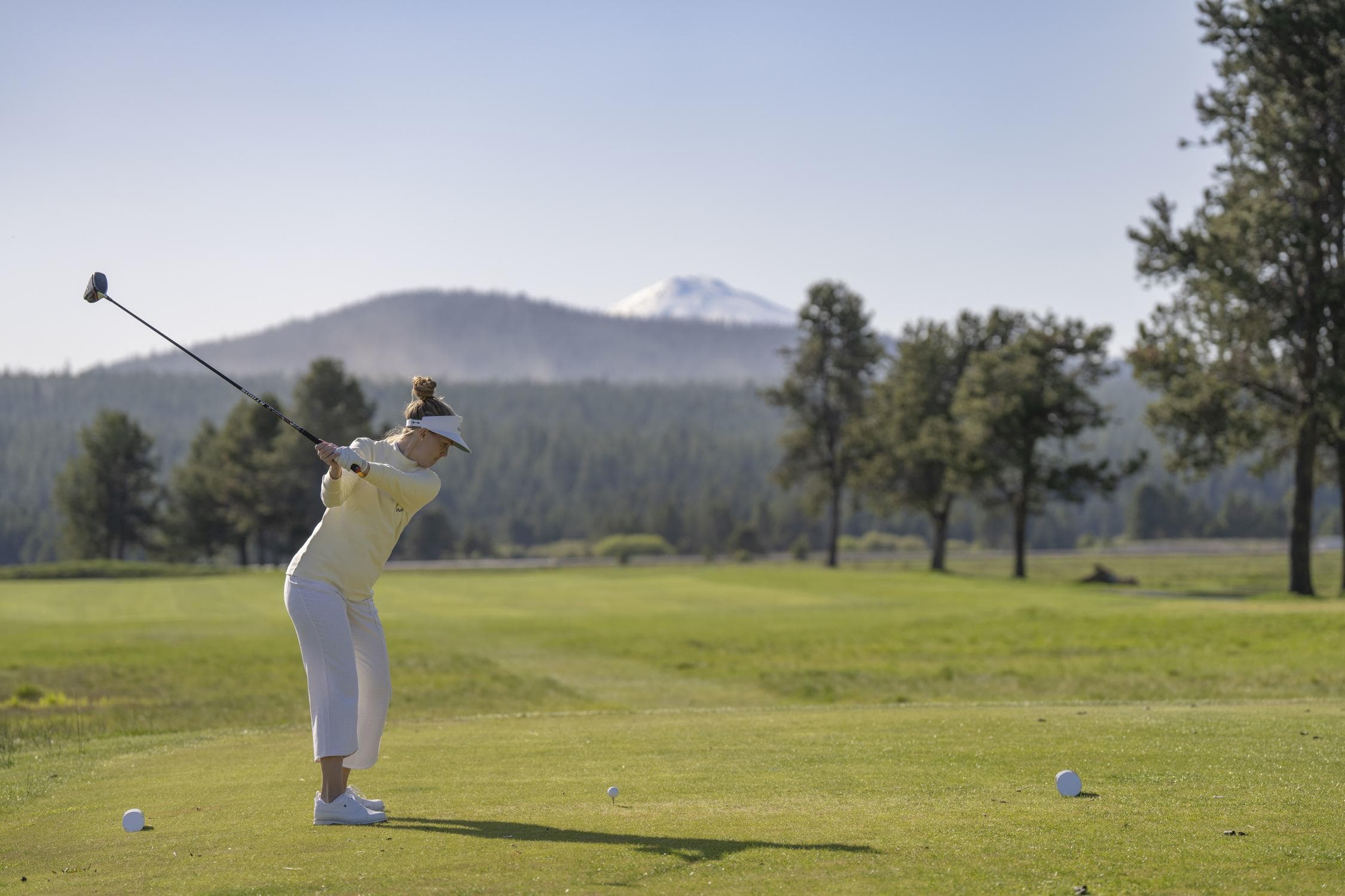A person in light clothing swings a golf club on a green golf course with trees and a mountain in the background on a clear day.