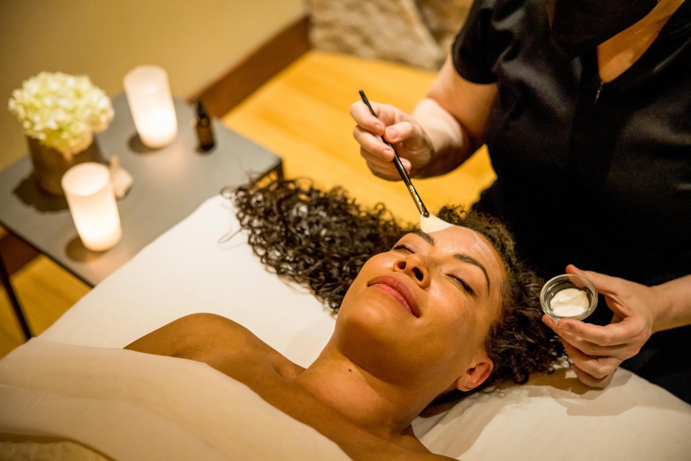 A person lies on a spa bed with eyes closed as an esthetician applies a facial mask with a brush. Lit candles and flowers are on a nearby table, creating a relaxing atmosphere.