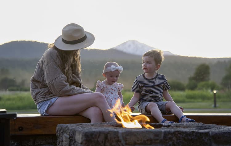 A woman in a hat sits with two young children by an outdoor fire pit, with green fields and a snow-capped mountain in the background.