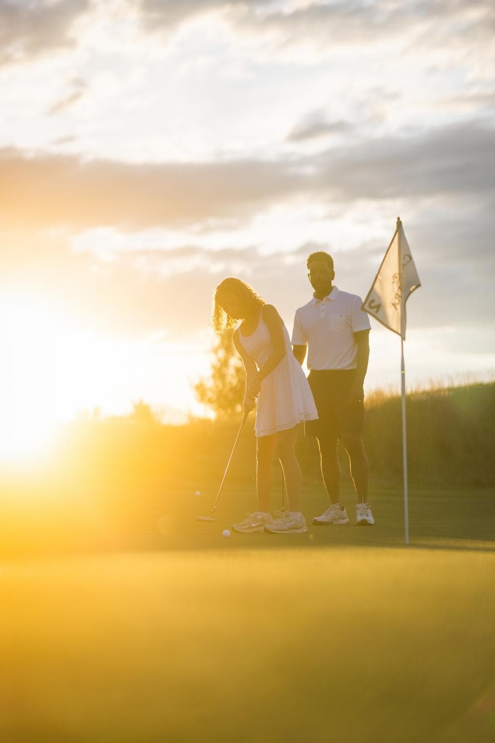 Two people playing golf at sunset; one is lining up a putt near the hole and flag while the other stands nearby, both dressed in white sports attire, with sunlight streaming in the background.