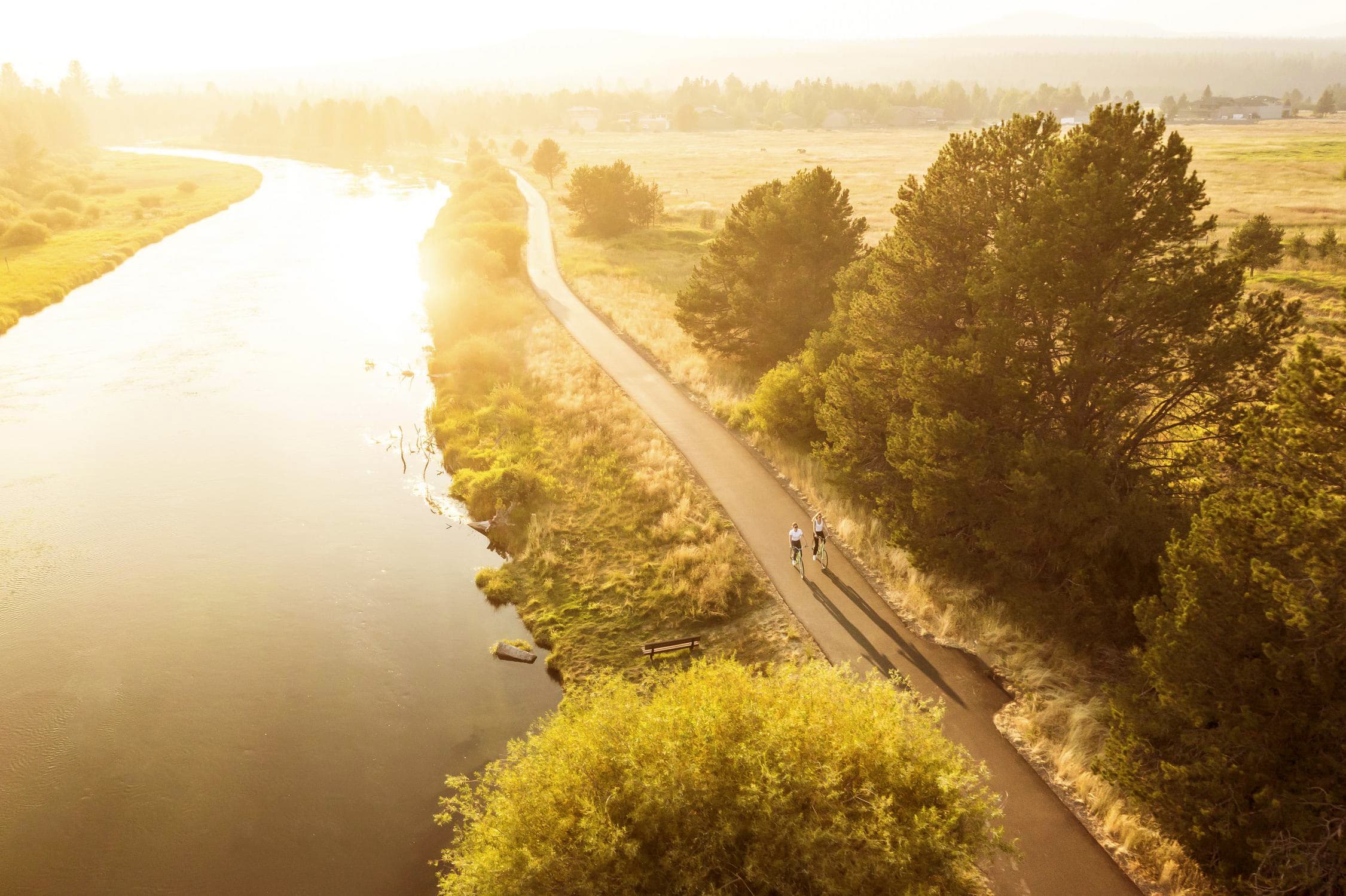 Aerial view of two people biking on a path beside a river at sunrise, with trees and grassy fields on both sides, and golden sunlight illuminating the scene.