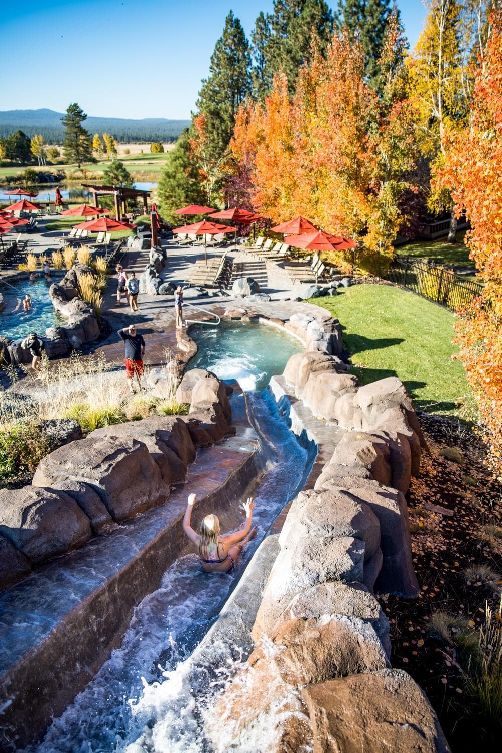 A person slides down a winding water slide at the Cove Aquatic Center surrounded by rocks, with people, pools, red umbrellas, and autumn trees in the background.