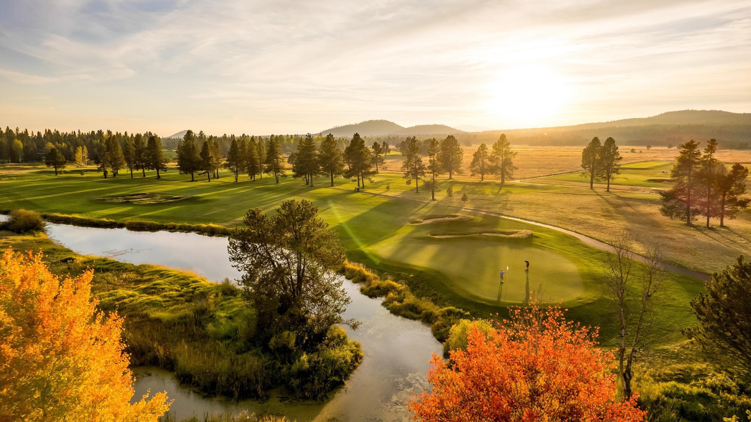 A scenic golf course at sunset, with manicured greens, trees, a winding waterway, and autumn foliage in the foreground. The sun casts a warm golden light over the landscape.