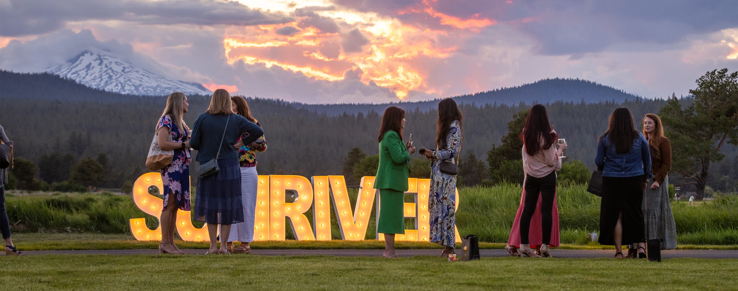 A group of women talking outdoors at sunset with mountains in the background and large illuminated letters spelling SURVIVE behind them.