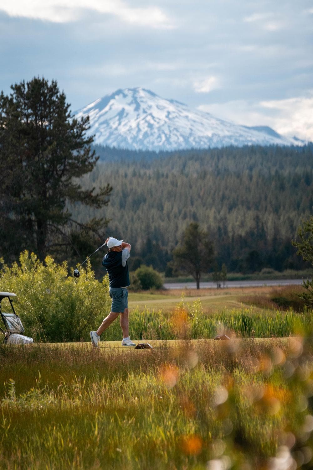 A golfer swings a club on a lush course, with tall trees and a snow-capped mountain in the background under a partly cloudy sky.
