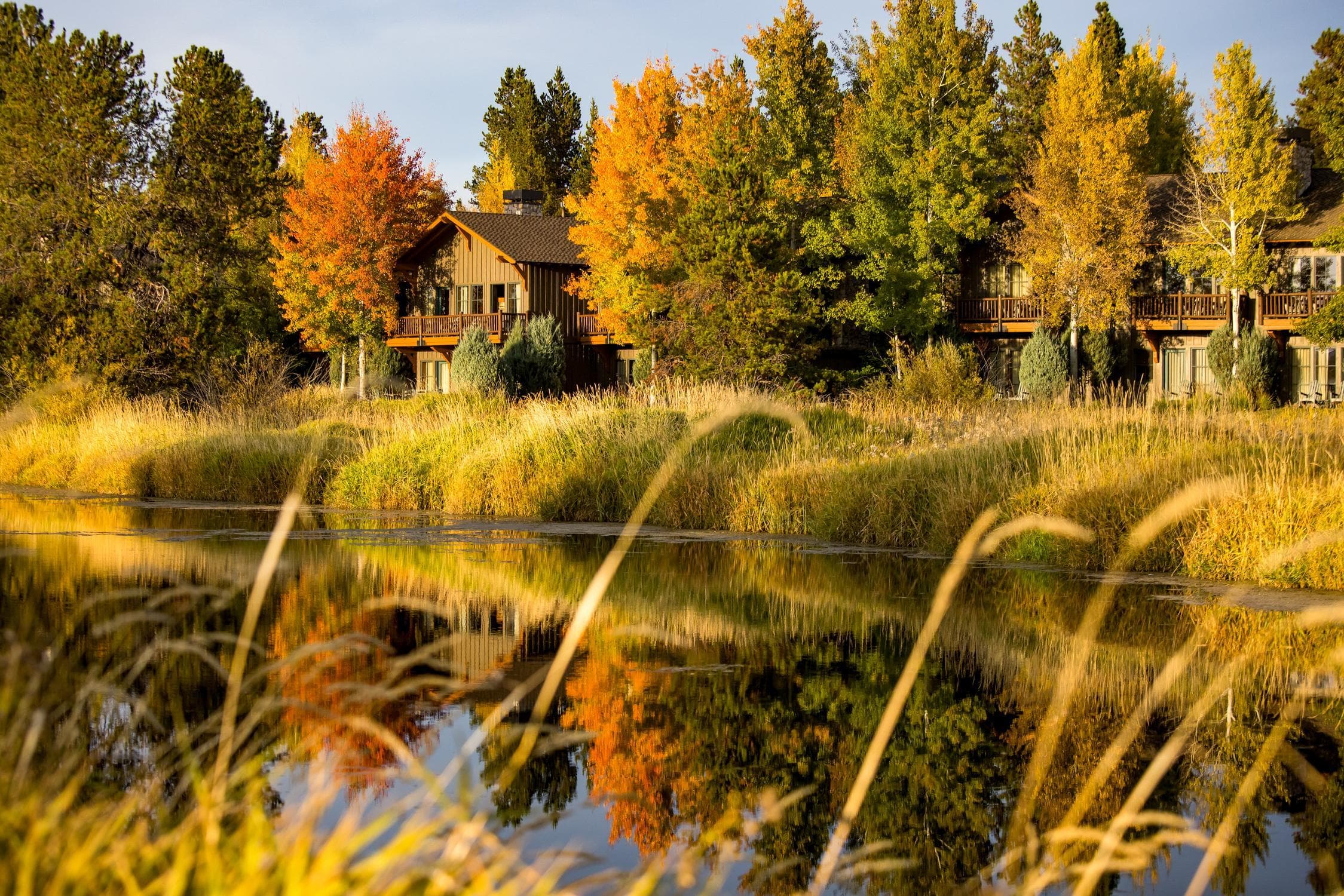 A wooden cabin surrounded by colorful autumn trees sits by a calm lake, with reflections of the foliage and grass visible in the water under clear skies.