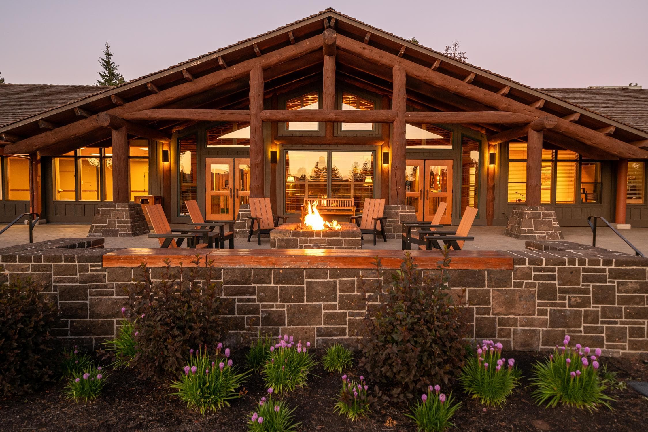 A lodge-style building with large wooden beams, a central fire pit surrounded by Adirondack chairs, stone wall, and blooming purple flowers in front at sunset.