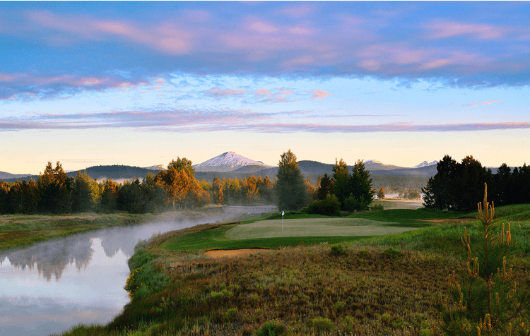 A golf course sits beside a calm river, surrounded by trees and grasses, with snow-capped mountains and a colorful sunrise sky in the background.