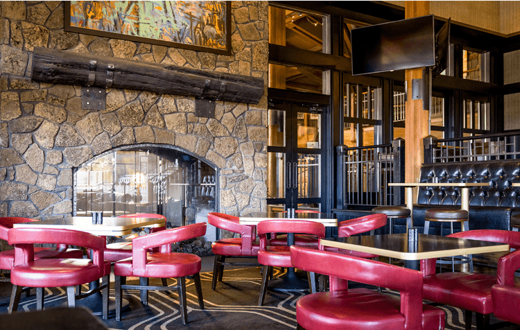 Cozy restaurant interior with red chairs, black tables, stone fireplace, TV, and artwork on the wall. Large windows and wooden accents create a warm, inviting atmosphere.