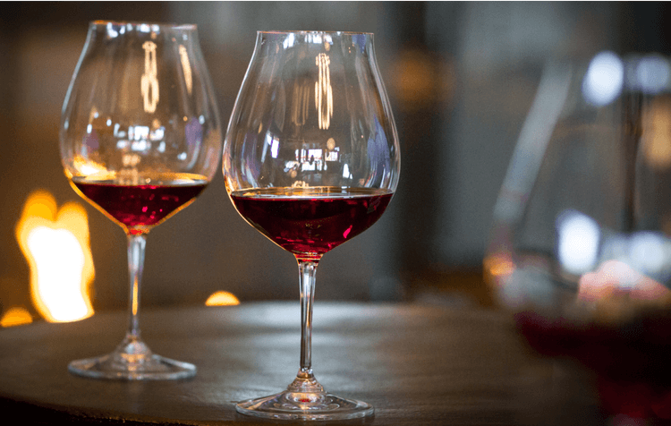 Two glasses of red wine sit on a round table with a soft, warm light and blurred fire in the background, creating a cozy atmosphere.