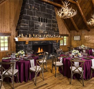 A rustic lodge dining room with round tables covered in purple cloths, floral centerpieces, wooden chairs, a stone fireplace with a fire, and antler chandeliers hanging from a wooden ceiling.