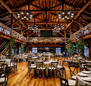Rustic wooden banquet hall with round tables set for an event, wooden chairs, chandeliers, exposed beams, and a balcony. Greenery decorates the space, creating a warm, inviting atmosphere.