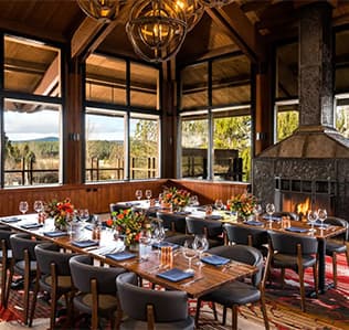 Elegant dining room with two long tables set with flowers and glassware, featuring large windows, a modern fireplace, wood beams, and views of trees outside. Warm, cozy atmosphere with natural light and contemporary decor.