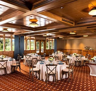 Elegant banquet room with round tables set for an event, featuring white tablecloths, floral centerpieces, wooden chairs, large windows, wood-paneled ceiling, and patterned carpet. Natural light brightens the space.