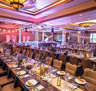 Elegant banquet hall set for a formal event, with long tables covered in blue tablecloths, neatly arranged plates, glasses, and silverware, decorative centerpieces, and string lights overhead.