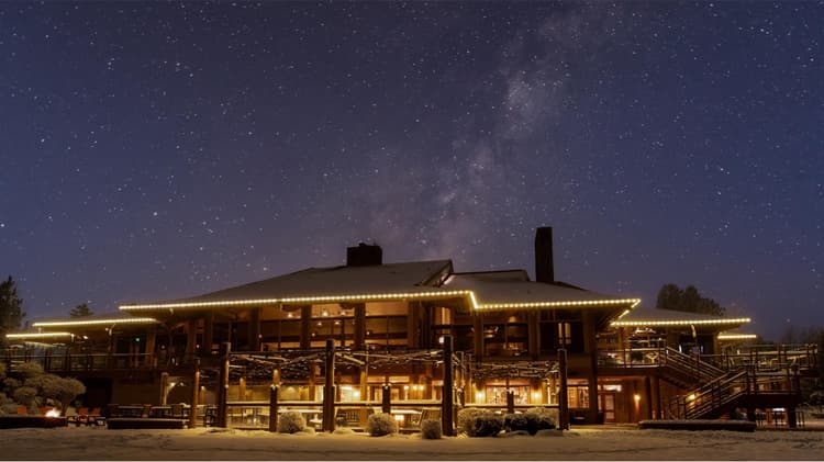 A large, warmly lit wooden lodge at night, surrounded by snow, stands under a star-filled sky with the Milky Way visible above.