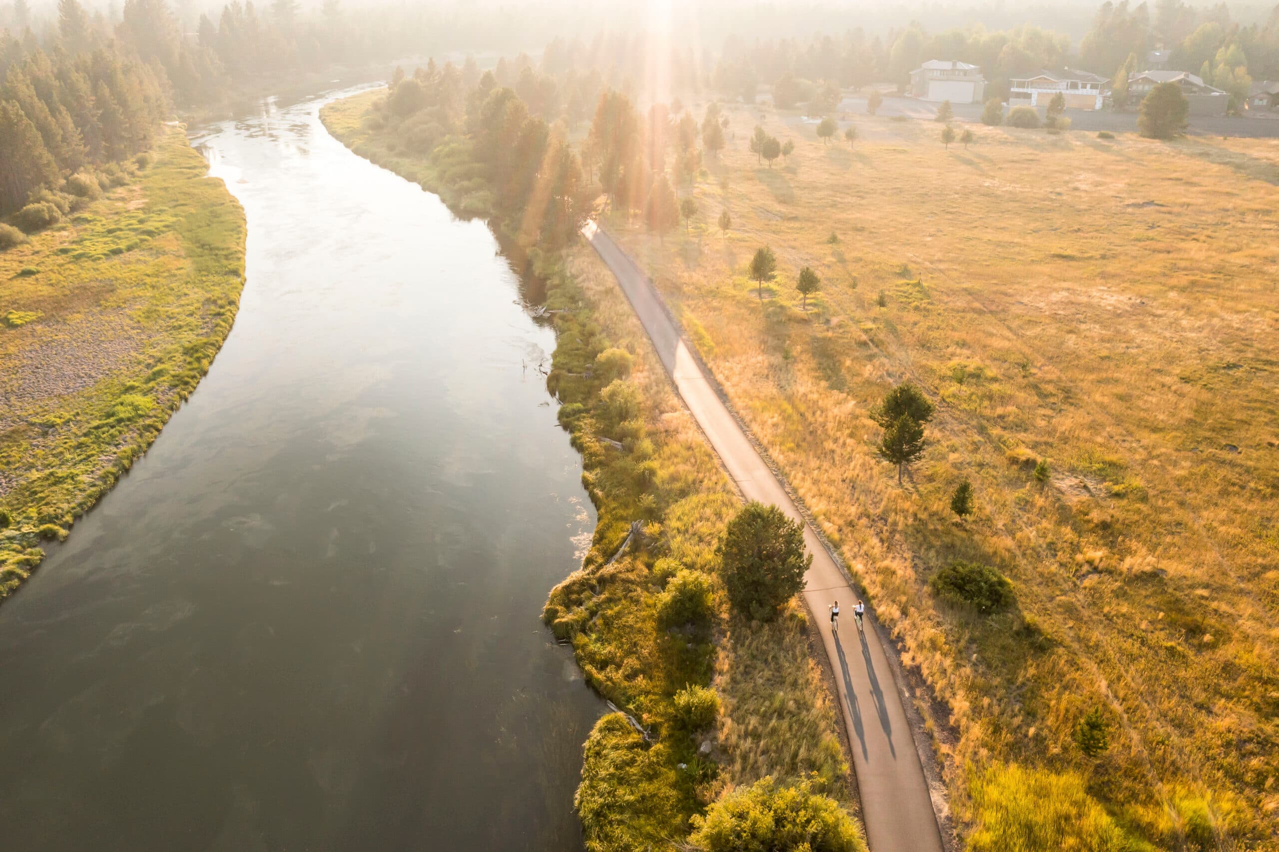 Aerial view of a river winding alongside a paved path where two cyclists ride through a sunlit, grassy landscape with trees and distant buildings.