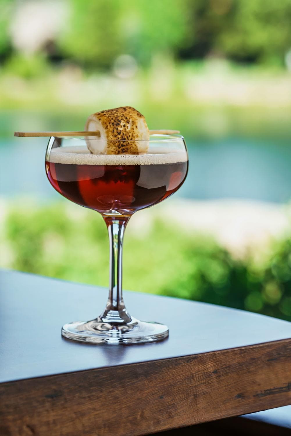 A dark cocktail in a coupe glass topped with a layer of foam and garnished with a toasted marshmallow on a skewer, set on a wooden table outdoors with a blurred nature background.