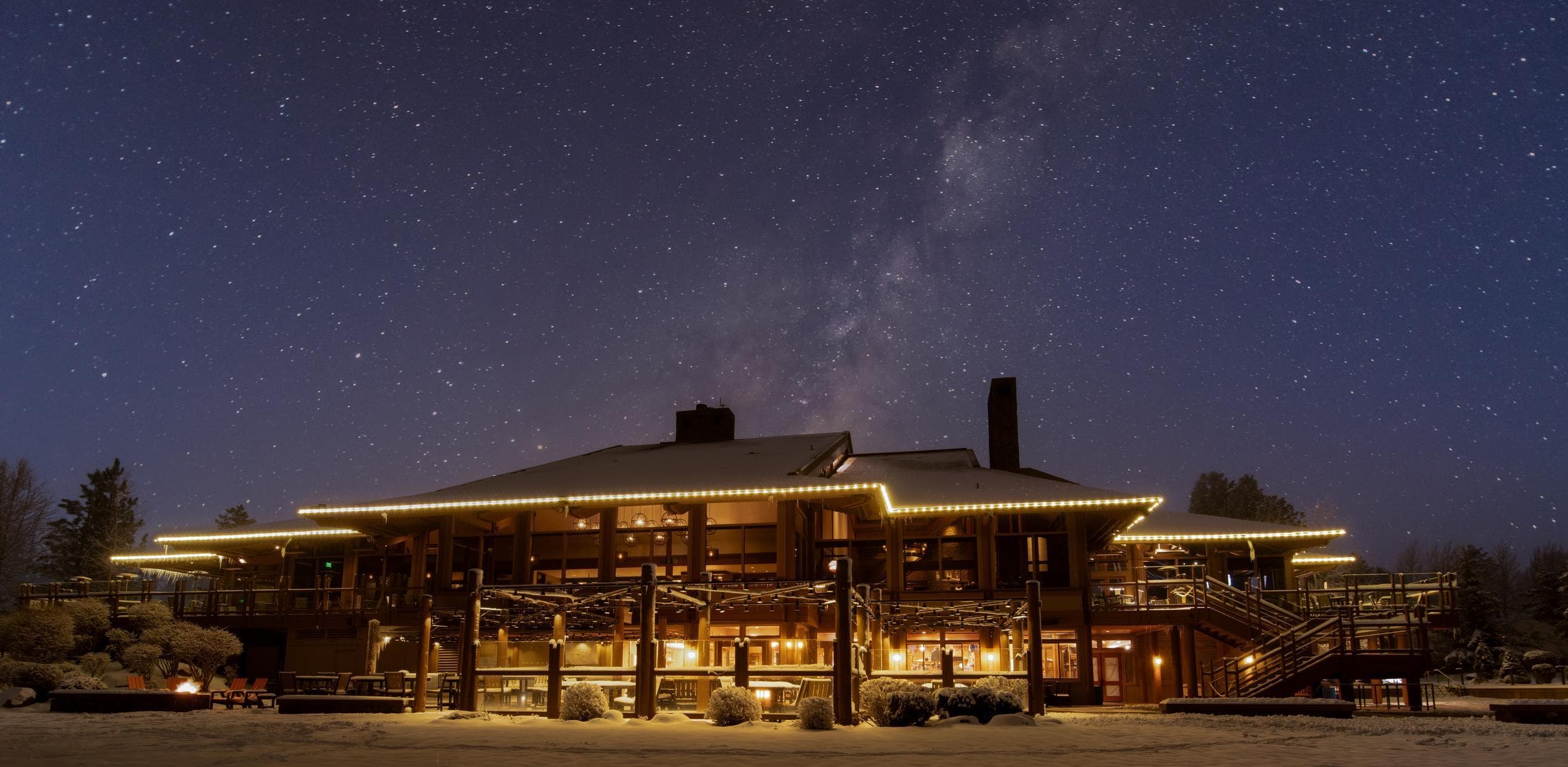 A large, warmly lit lodge with snow on the ground sits under a clear night sky filled with stars and the Milky Way.