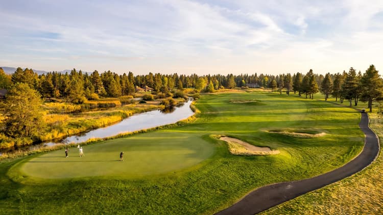 Aerial view of a golf course with three people on a green, a sand trap nearby, a winding path, and a river bordered by trees under a partly cloudy sky.