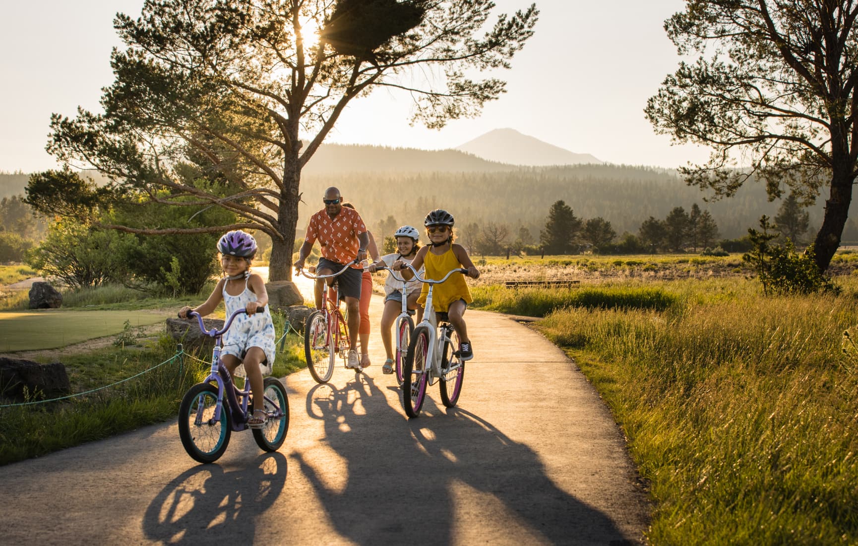 A family rides bicycles together along a paved path at sunset, with children wearing helmets and a parent riding behind them, surrounded by pine trees and mountain views.