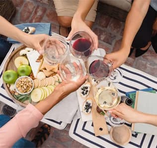 Five people toast with glasses of wine over a table with apples, cheese, crackers, and snacks, creating a festive and social picnic atmosphere.